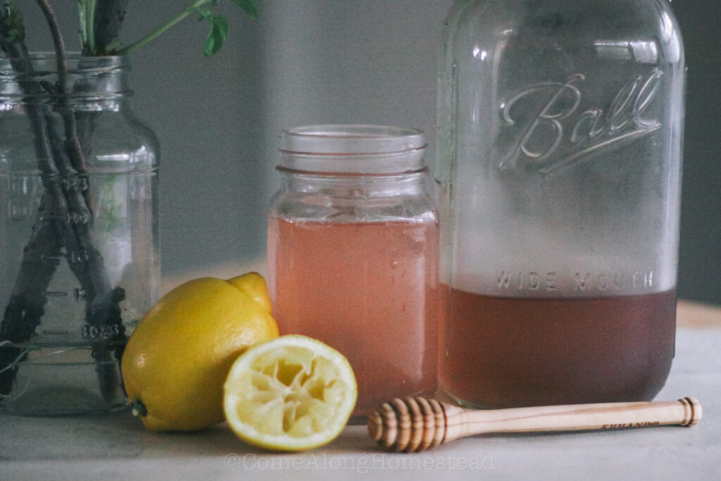 glass of pink lemonade with a cut up lemon and a honey dipper on the table in front of it.