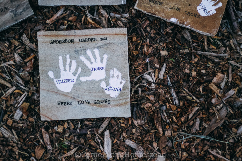 square stepping stone with three kid handprints in white paint saying the phrase "anderson garden. where love grows"
