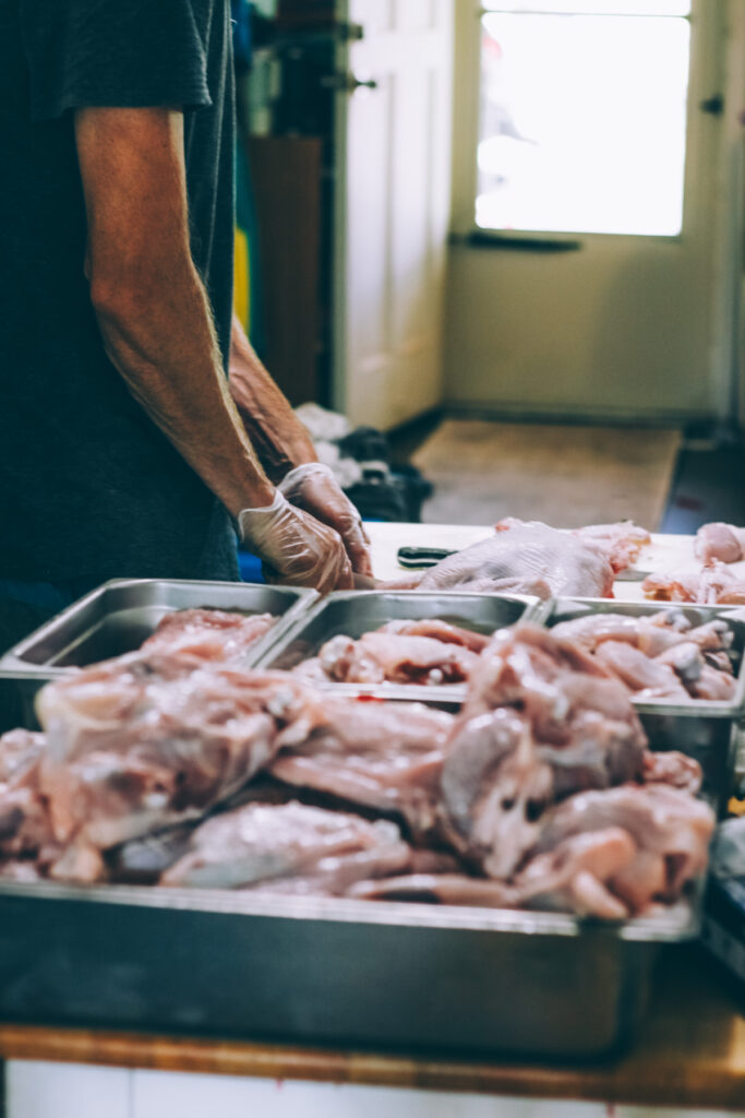 Different sizes of bins containing various cuts of chicken, with hands cutting a chicken in the background.